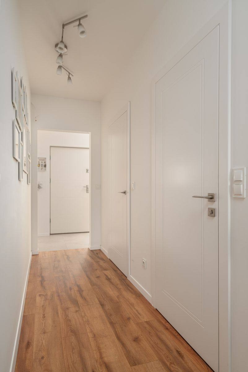 Jasny korytarz z drewnianą podłogą, białymi drzwiami z ozdobnymi ramkami i nowoczesnym oświetleniem sufitowym. Widoczne detale wykończenia i minimalistyczny design.