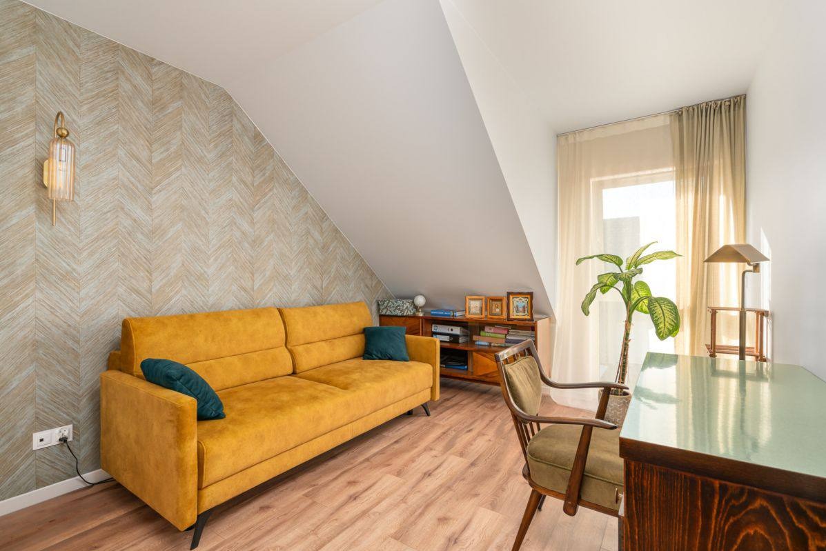 Pokój na poddaszu ze skośnym sufitem. Żółta sofa z poduszkami, drewniane biurko i krzesło. Na ścianie wzorzysta tapeta i lampa. Jasne drewniane panele na podłodze.