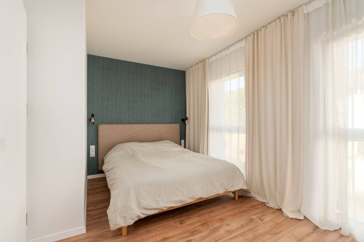 Jasna sypialnia z drewnianą podłogą, łóżkiem z beżowym zagłówkiem i zieloną ścianą z tapetą. Okno zasłonięte kremowymi, zwiewnymi zasłonami.