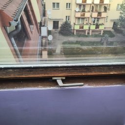 NAPRAWA OKIEN by Vitalii Luchkovskyi - Okno dachowe z widokiem na budynek mieszkalny. Drewniana rama okienna, uchwyt okna w pozycji zamkniętej. Ściana pomalowana na fioletowo. Widoczne ślady użytkowania.