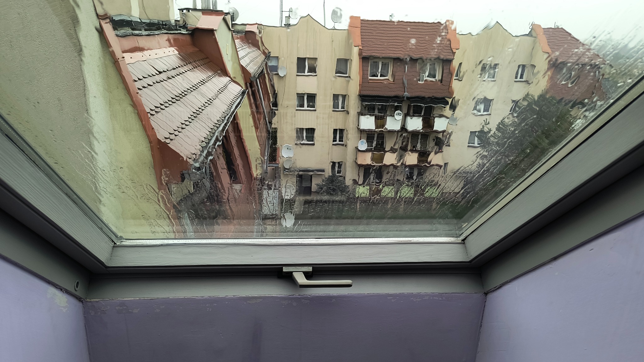 Widok z okna dachowego na budynki w deszczowy dzień. Krople deszczu zamazują obraz za szybą. Szara rama okna i fioletowa ściana w dolnej części kadru.