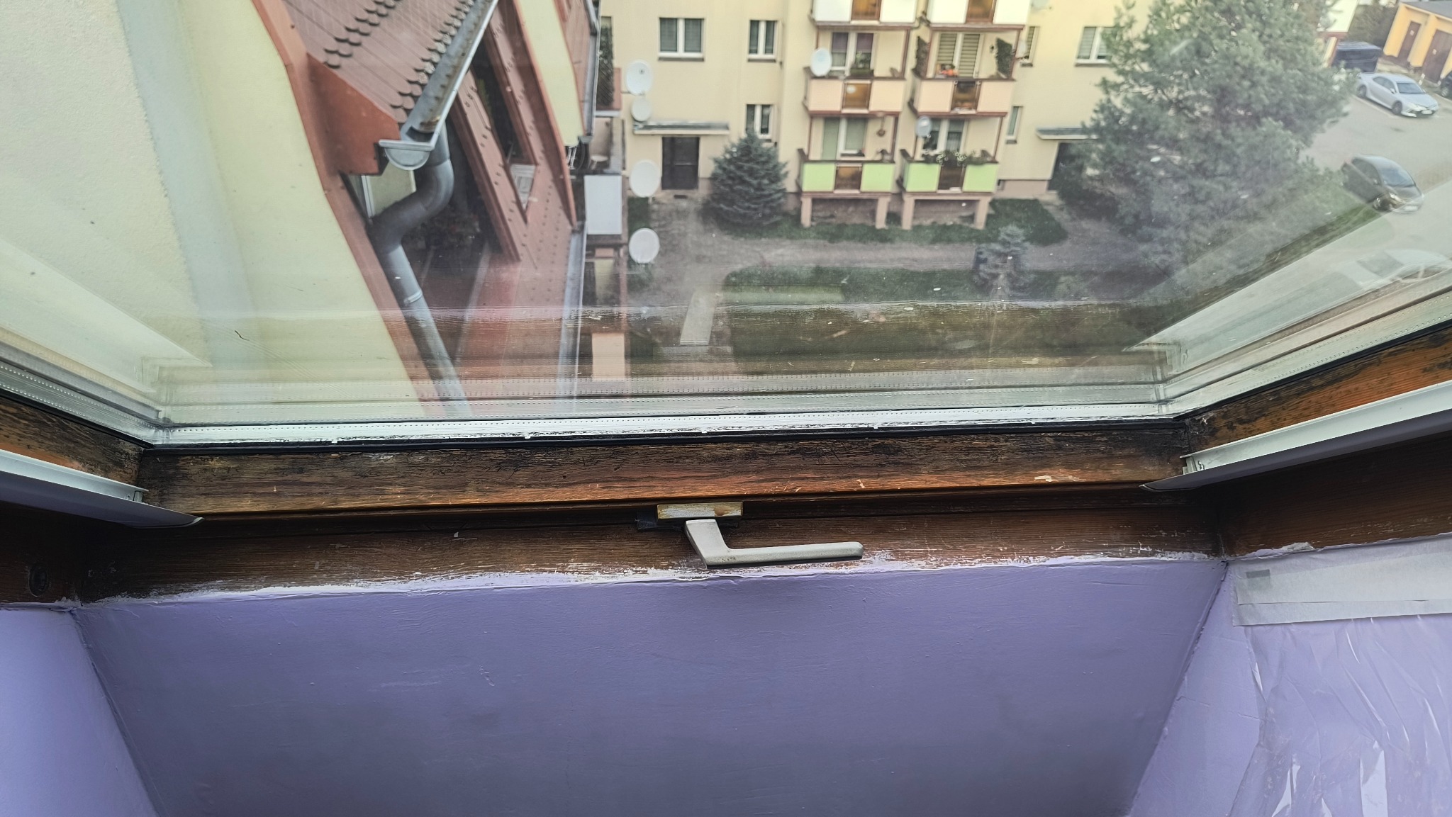 Okno dachowe z widokiem na budynek mieszkalny. Drewniana rama okienna, uchwyt okna w pozycji zamkniętej. Ściana pomalowana na fioletowo. Widoczne ślady użytkowania.