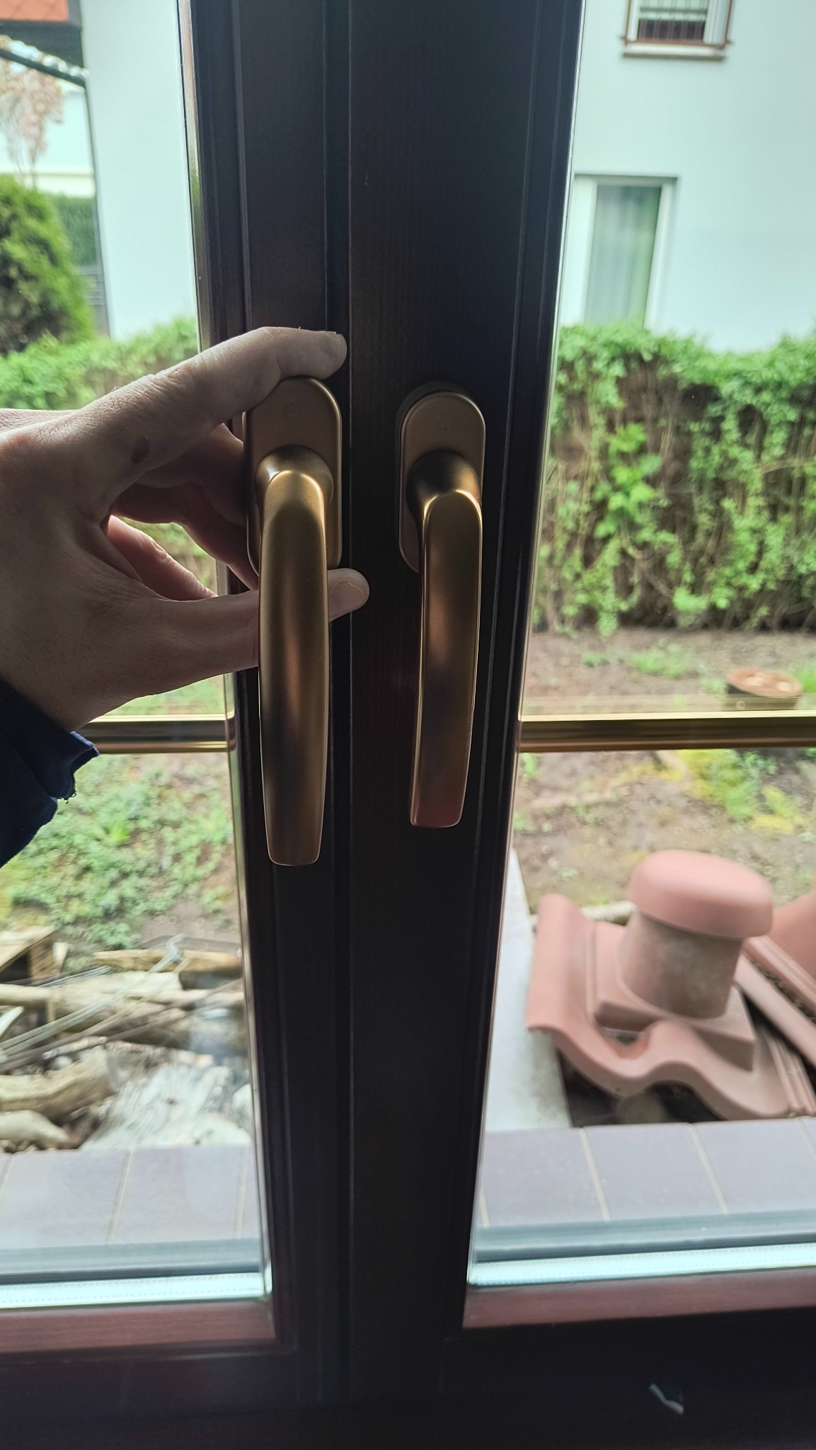 Brązowe okno z dwiema złotymi klamkami, otwarte przez dłoń. Widok na ogród z zielenią i fragmentem dachu. Detal stolarki okiennej.