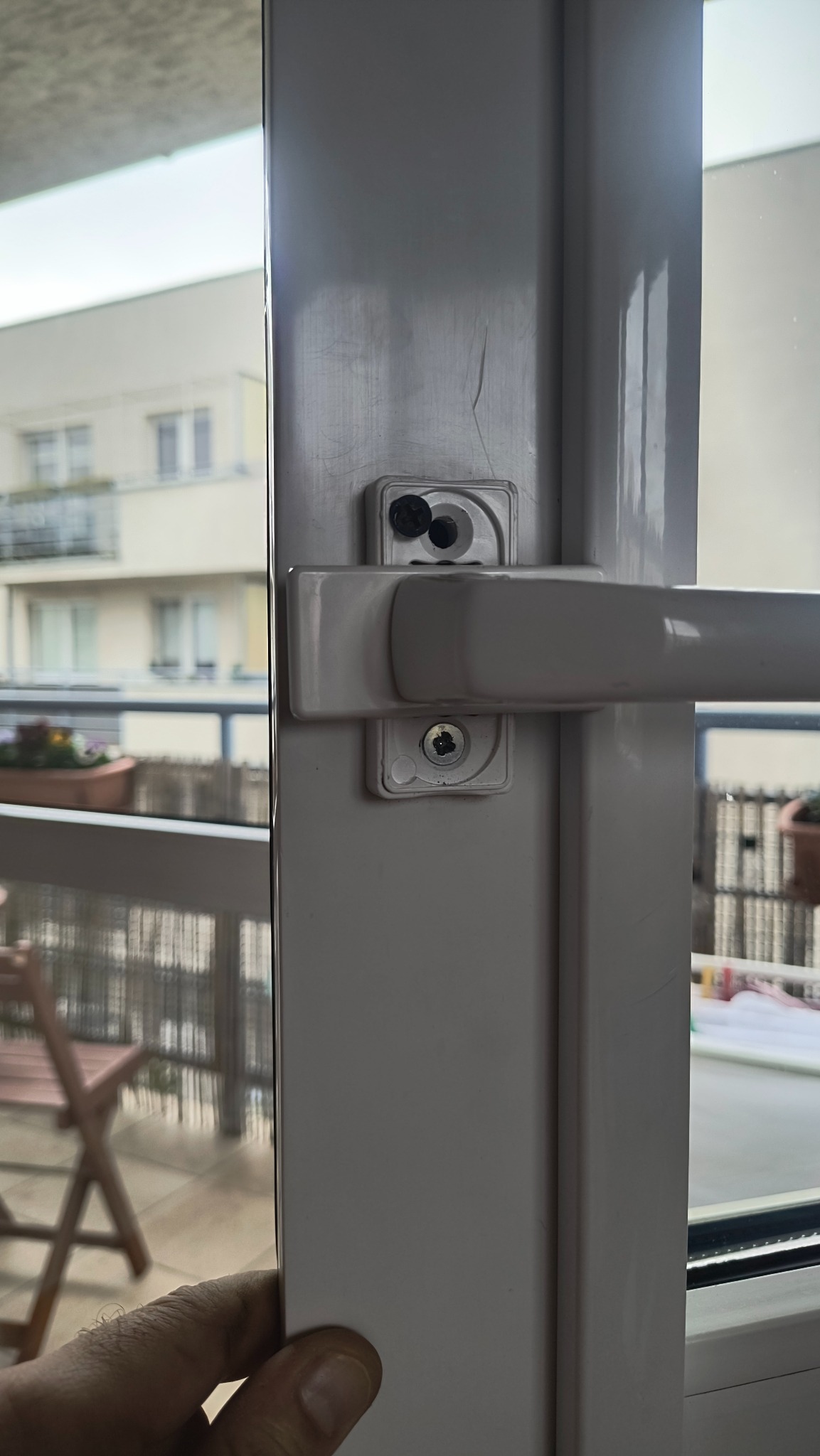 Białe okno PCV z widocznym mechanizmem blokującym i klamką, na tle balkonu z kwiatami i budynkiem w oddali. Detal montażu okna.