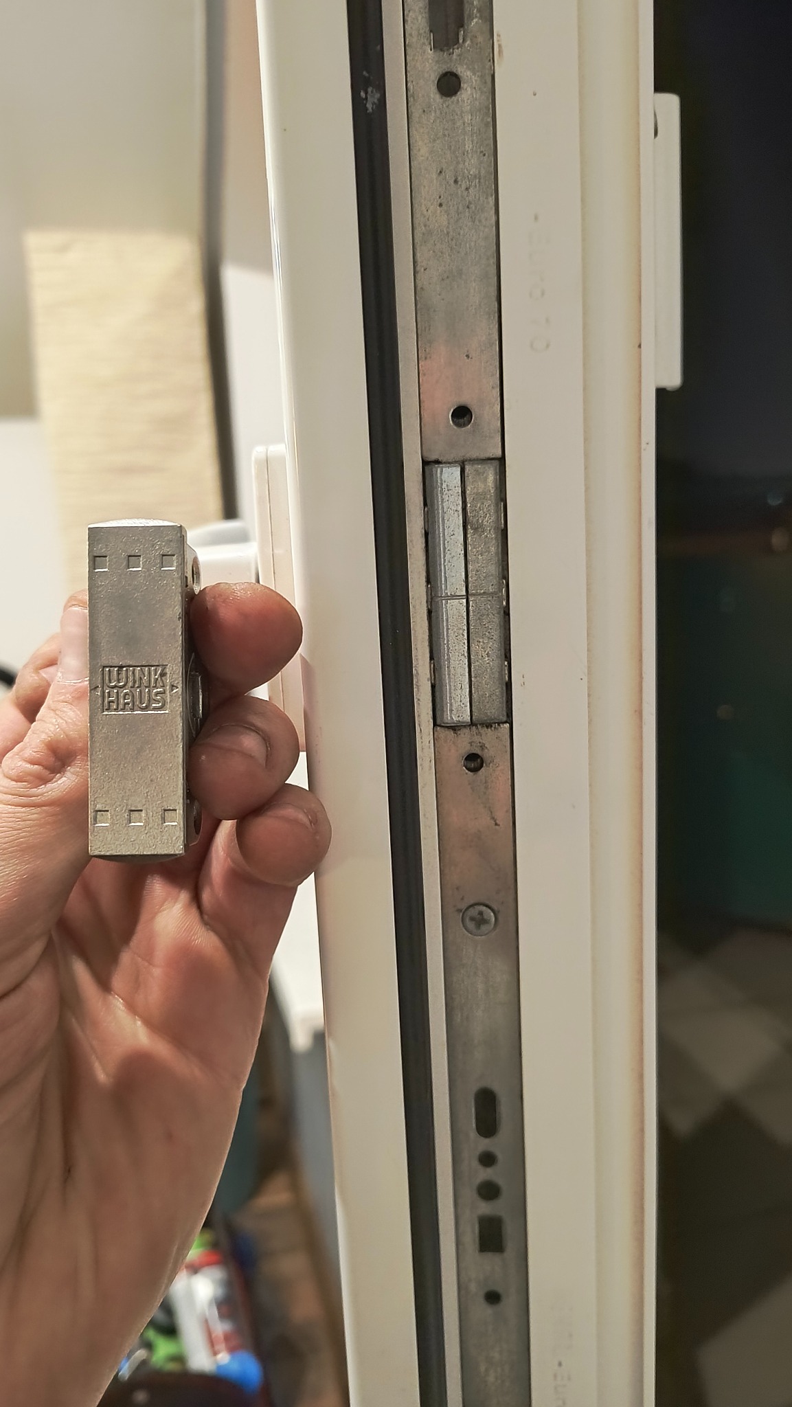 Regulacja okna: dłoń przytrzymuje element zamka Winkhaus przy białej ramie okiennej z widoczną listwą zamykającą. Detal mechanizmu okiennego.