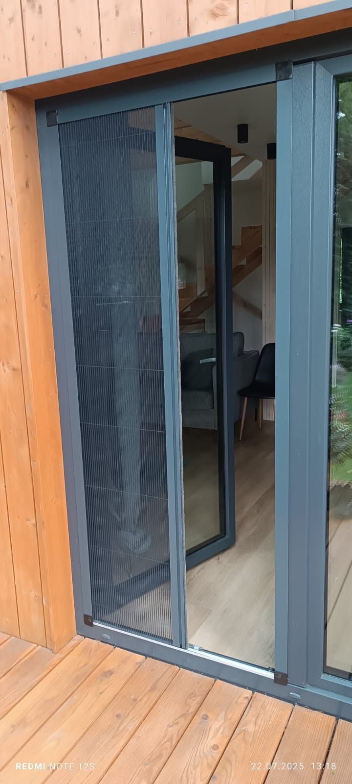 Szare przesuwne drzwi balkonowe z moskitierą plisowaną, widok z zewnątrz na drewniany taras i wnętrze domu z schodami. Nowoczesne rozwiązanie.