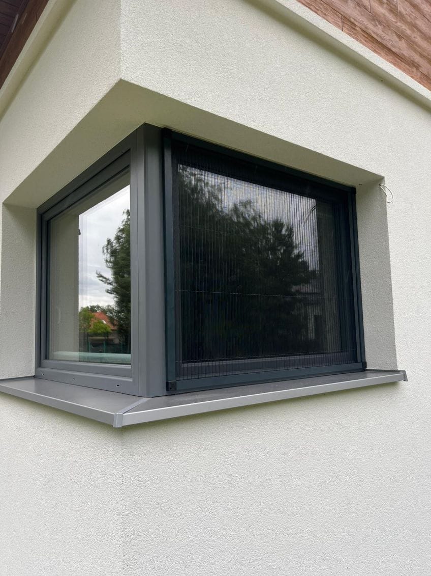 Szare okno PCV z moskitierą plisowaną, zamontowane w białej elewacji budynku. Widok z zewnątrz, z bliska, z widocznym odbiciem drzewa w szybie.