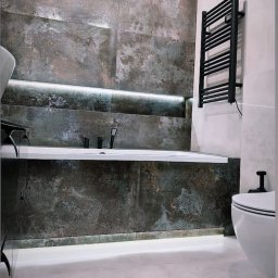 S.H. REMONTY - Nowoczesna łazienka z wanną obudowaną płytkami imitującymi rdzę, podświetleniem LED i czarnym grzejnikiem drabinkowym. Minimalistyczny design, widoczny fragment toalety.