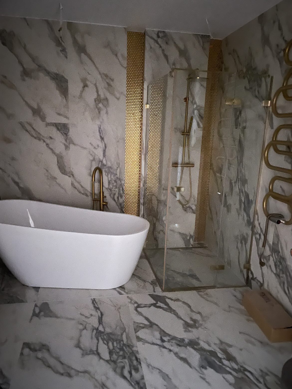 Elegancka łazienka z wanną wolnostojącą, kabiną prysznicową ze złotymi detalami i marmurowymi płytkami na ścianach i podłodze. Nowoczesny design z akcentami luksusu.