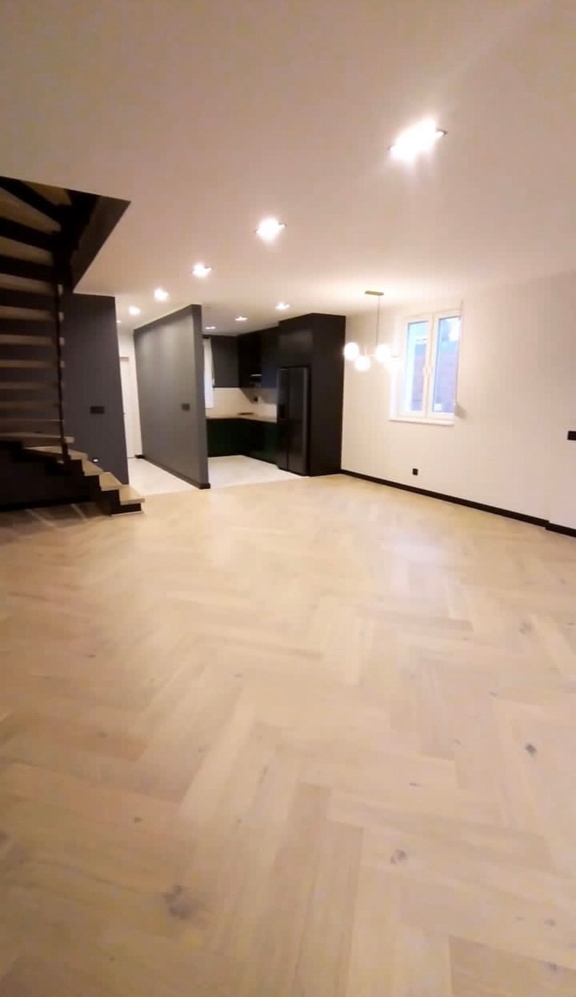 Jasna podłoga w jodełkę, ciemne schody i kuchnia w otwartej przestrzeni. Nowoczesne oświetlenie i minimalistyczny design. Widok na okno.