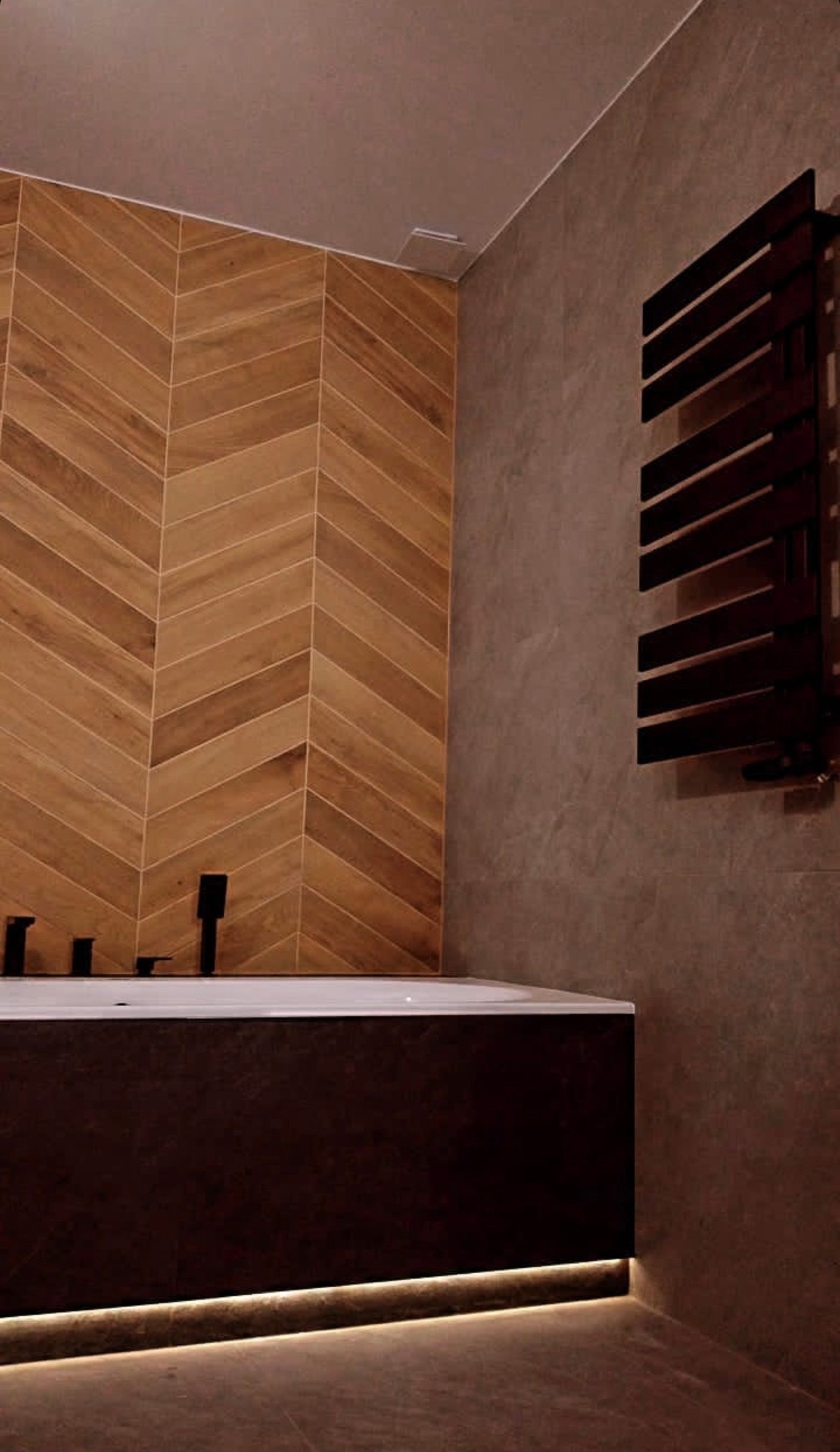 Nowoczesna łazienka z drewnopodobną glazurą ułożoną w jodełkę, czarną armaturą i grzejnikiem drabinkowym. Podświetlenie LED podkreśla obrys wanny.