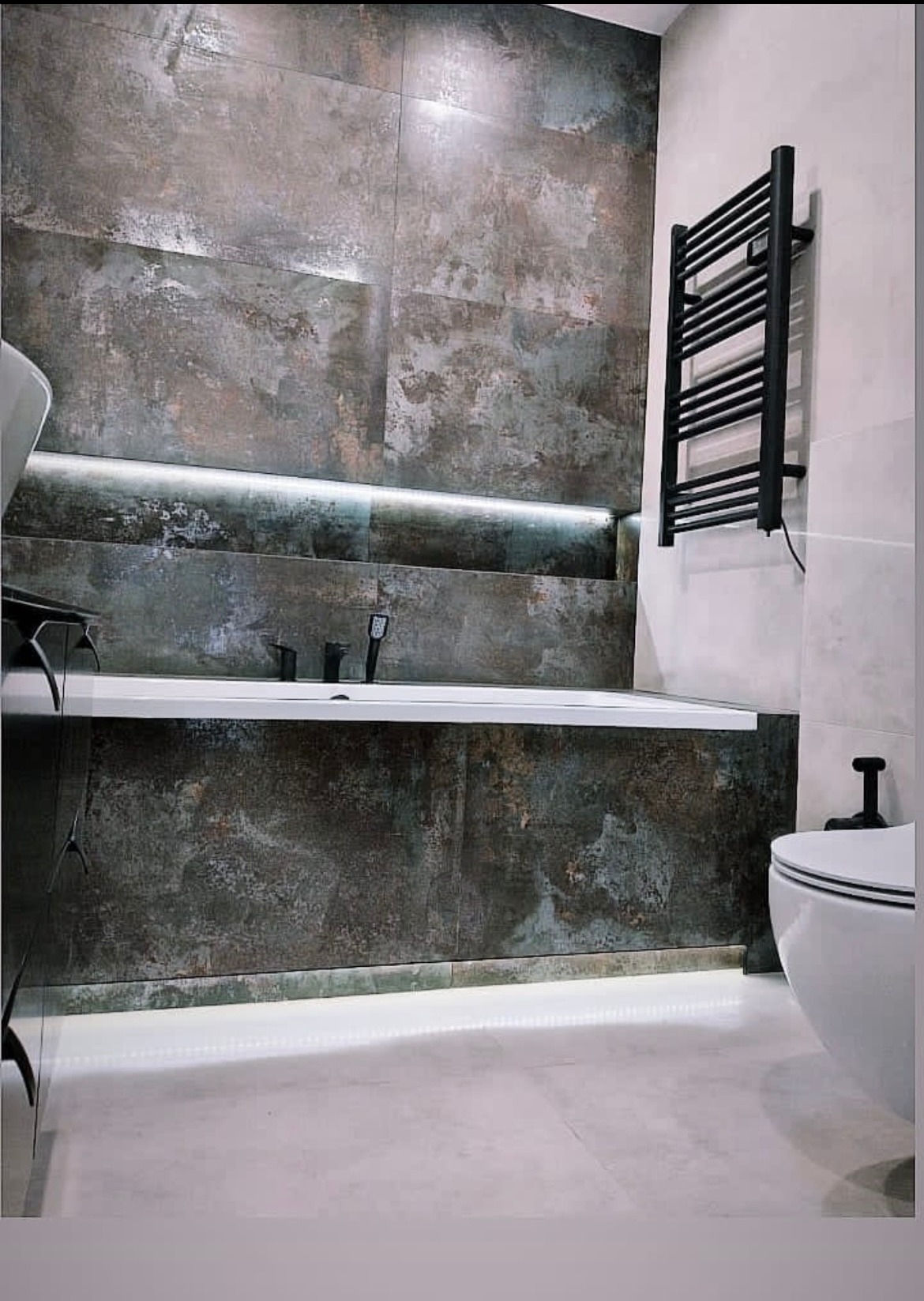 Nowoczesna łazienka z wanną obudowaną płytkami imitującymi rdzę, podświetleniem LED i czarnym grzejnikiem drabinkowym. Minimalistyczny design, widoczny fragment toalety.