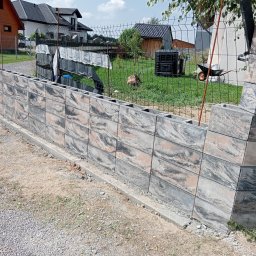 Firma Handlowo Usługowa Atut - Wznoszenie ogrodzenia z szarej, wzorzystej kostki betonowej. Widoczny fragment konstrukcji, siatka ogrodzeniowa w tle, elementy krajobrazu.