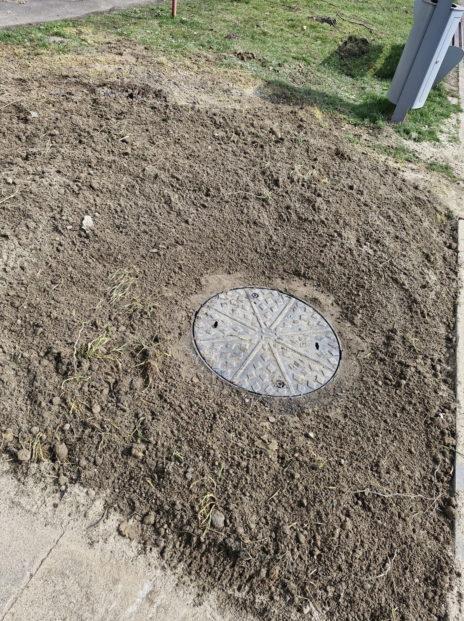 Wyrównany teren wokół studzienki kanalizacyjnej, świeża ziemia, widoczne ślady po pracach ziemnych. Fragment chodnika i trawnika w tle.