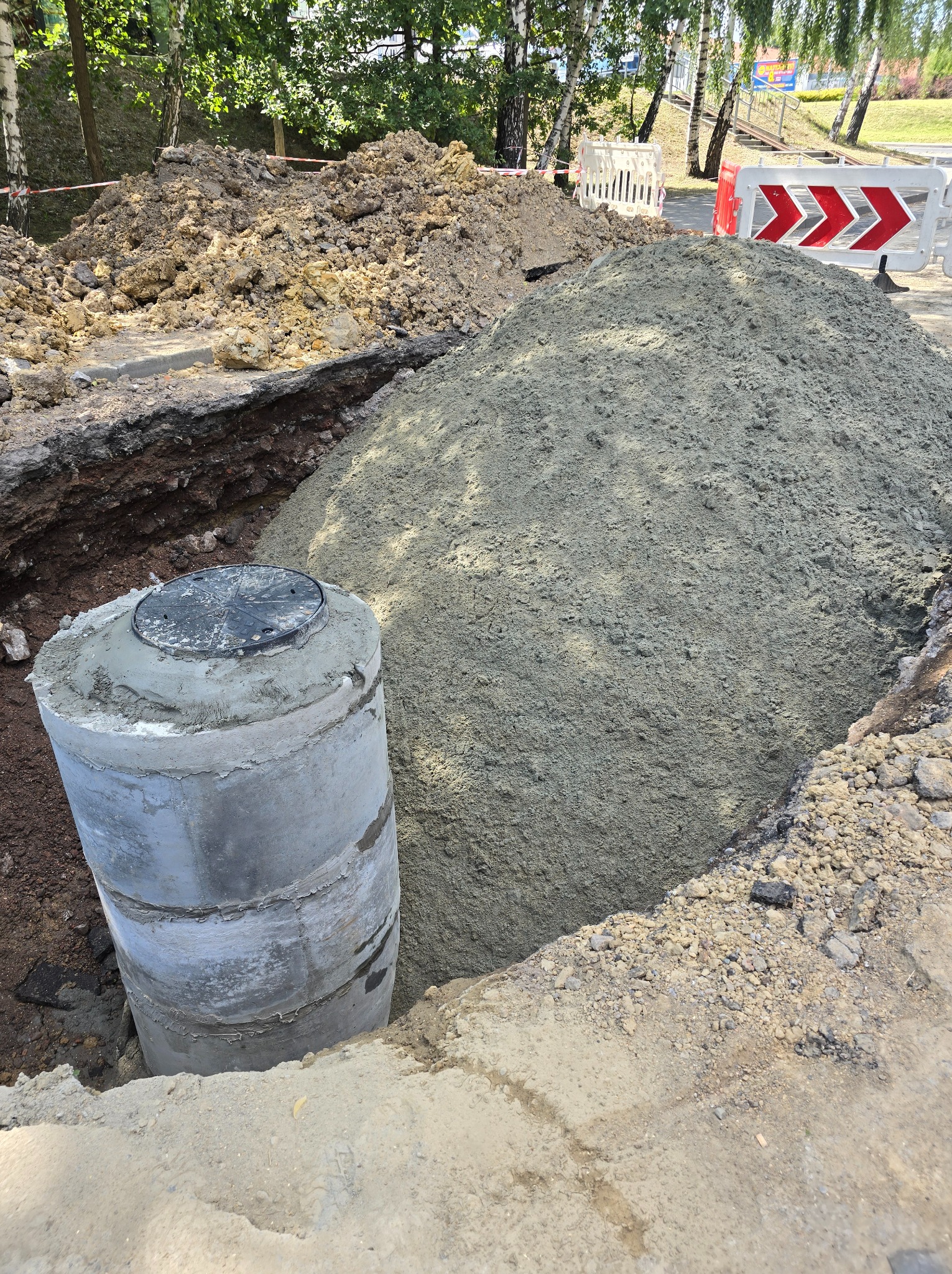 Betonowy krąg studzienny w wykopie, obok hałda piasku i ziemi. Widoczne zabezpieczenia wykopu i drzewa w tle. Prace ziemne przy budowie kanalizacji.