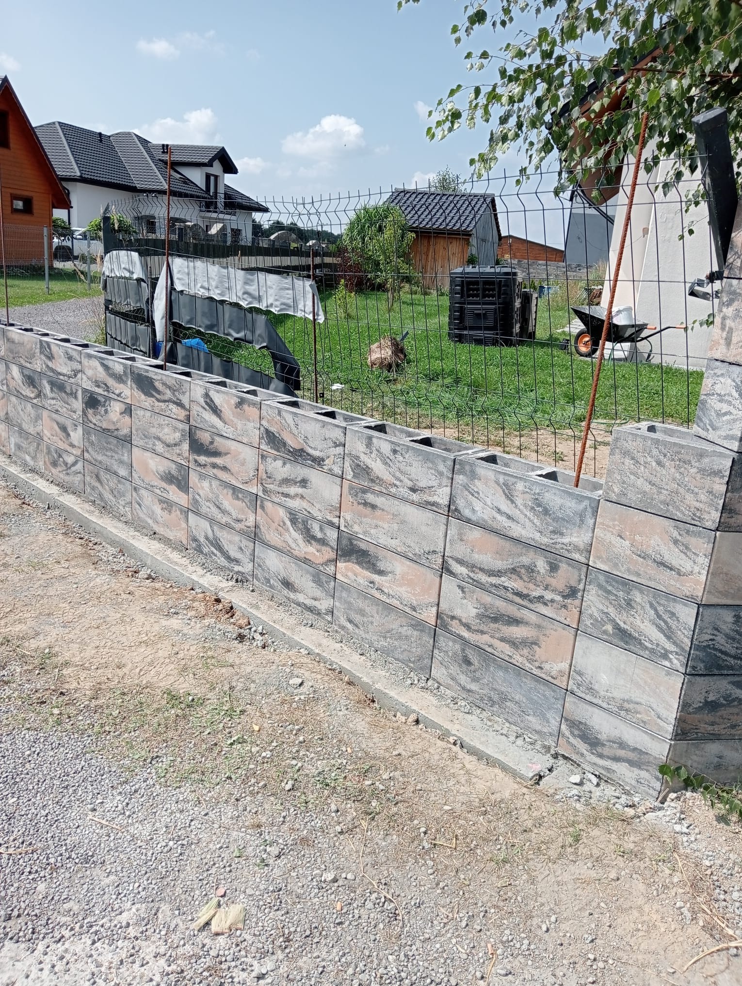 Wznoszenie ogrodzenia z szarej, wzorzystej kostki betonowej. Widoczny fragment konstrukcji, siatka ogrodzeniowa w tle, elementy krajobrazu.