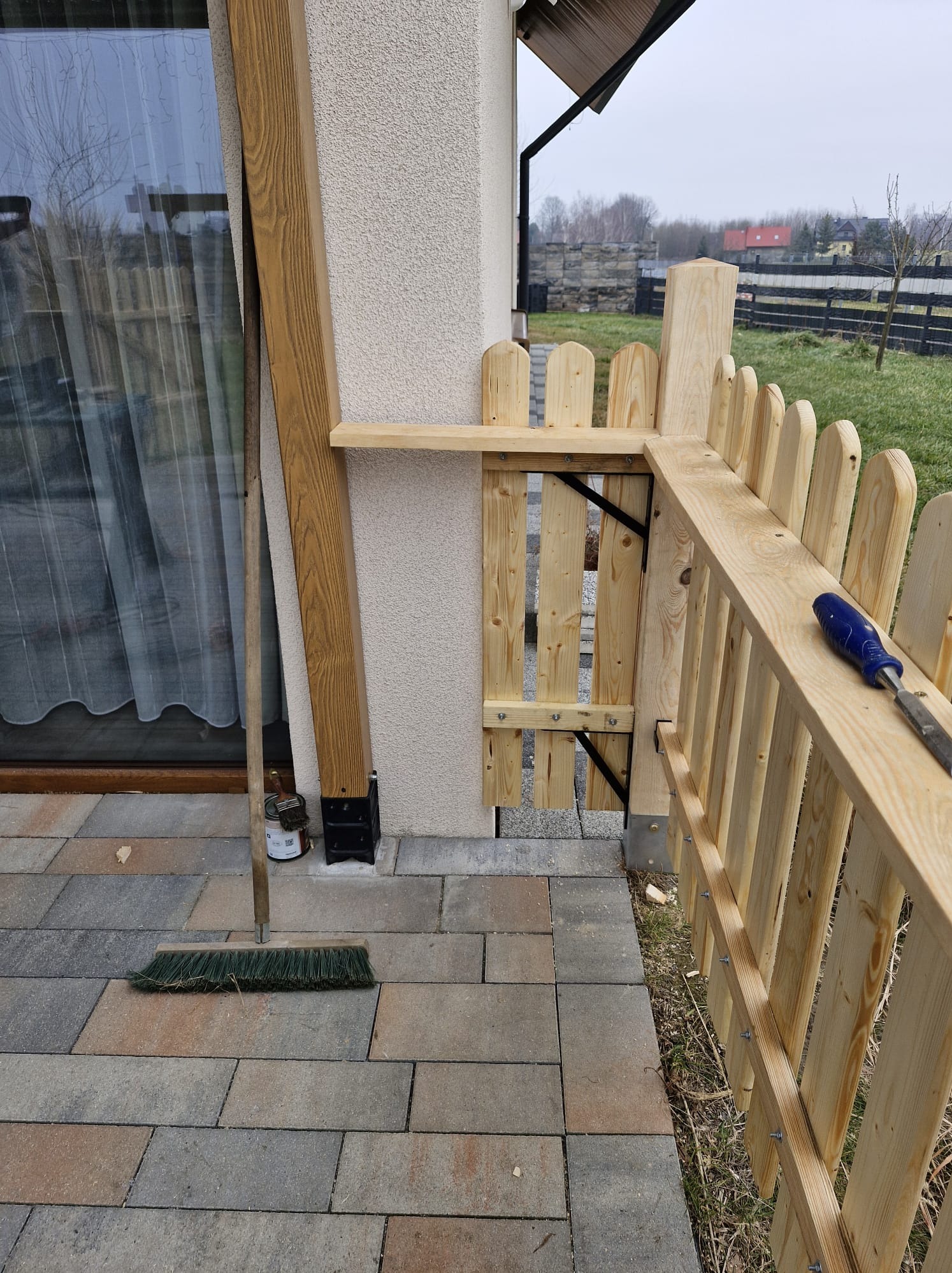 Fragment montowanego drewnianego ogrodzenia z furtką, widoczne narzędzia i kostka brukowa. Prace wykończeniowe przy domu.