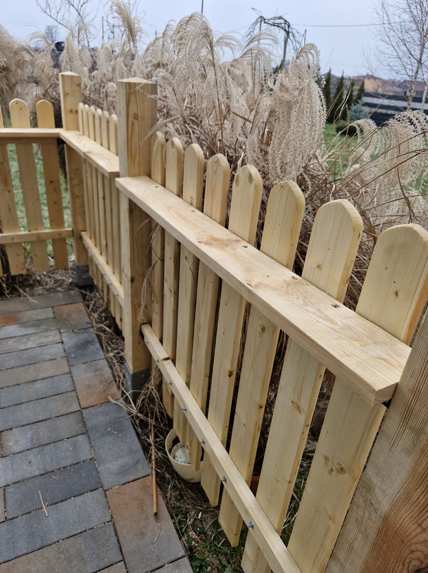 Jasne, drewniane ogrodzenie z palików, z widocznymi śrubami, oddzielające ścieżkę z kostki od ozdobnych traw. Ujęcie z bliska, podkreślające detale konstrukcji.