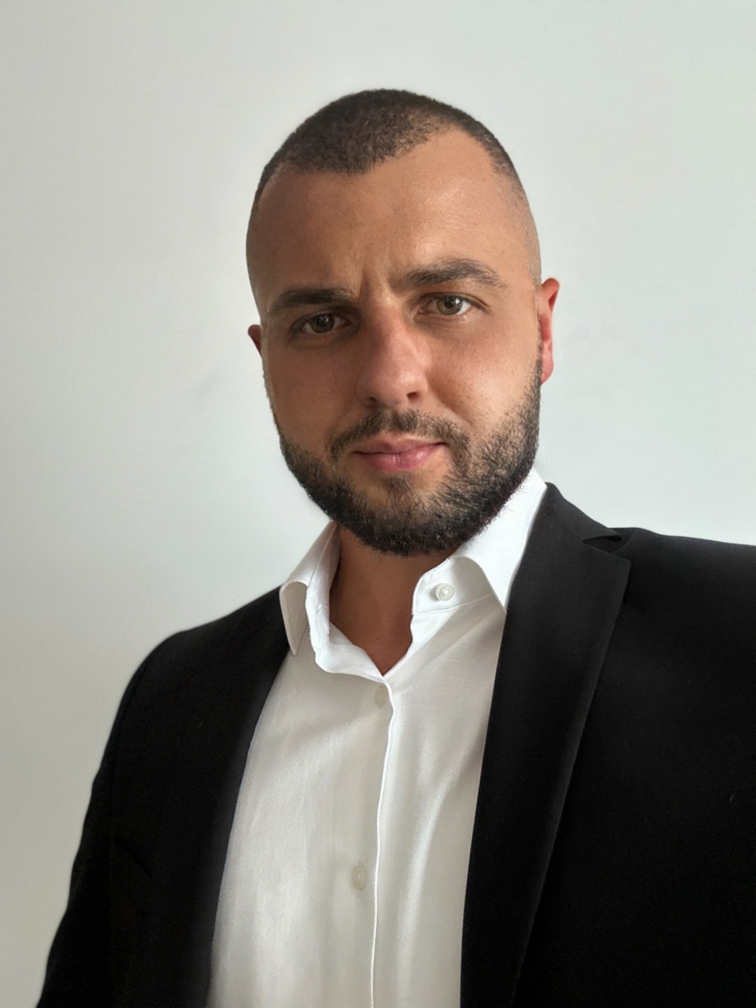 Portret mężczyzny z brodą w białej koszuli i czarnej marynarce na jasnym tle. Ujęcie od przodu, skupione na twarzy i górnej części ciała. Profesjonalny wygląd.