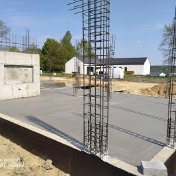 W.P.INVEST - Zalany fundament z wystającymi prętami zbrojeniowymi, widoczny betonowy element konstrukcyjny. W tle budynek z panelami słonecznymi i przenośna toaleta.