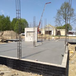 W.P.INVEST - Wylany fundament z wystającymi prętami zbrojeniowymi na placu budowy domu jednorodzinnego. W tle widoczny dźwig i częściowo zadaszona konstrukcja budynku.