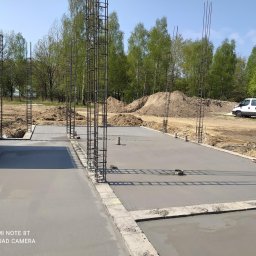 W.P.INVEST - Zalany fundament z wystającymi zbrojeniami pionowymi na budowie domu jednorodzinnego. W tle hałda ziemi i biały samochód dostawczy.