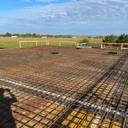 W.P.INVEST - Zbrojenie płyty fundamentowej z widocznym szalunkiem i wystającymi prętami, na tle krajobrazu z polami i błękitnym niebem. Cień osoby w lewym dolnym rogu.