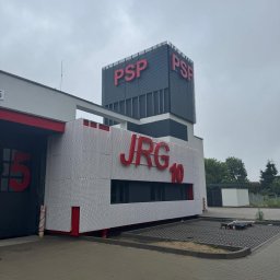 W.P.INVEST - Nowoczesny budynek straży pożarnej z czerwonymi napisami PSP i JRG 10 na tle szarego nieba. Charakterystyczna elewacja z perforowanej blachy.