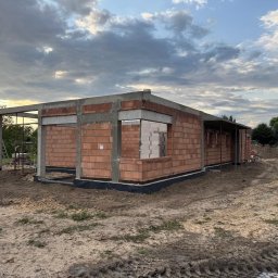 W.P.INVEST - Surowy stan domu z czerwonej cegły i betonu pod pochmurnym niebem. Widoczne zbrojenia i pustaki. Etap budowy z widocznymi elementami konstrukcyjnymi.