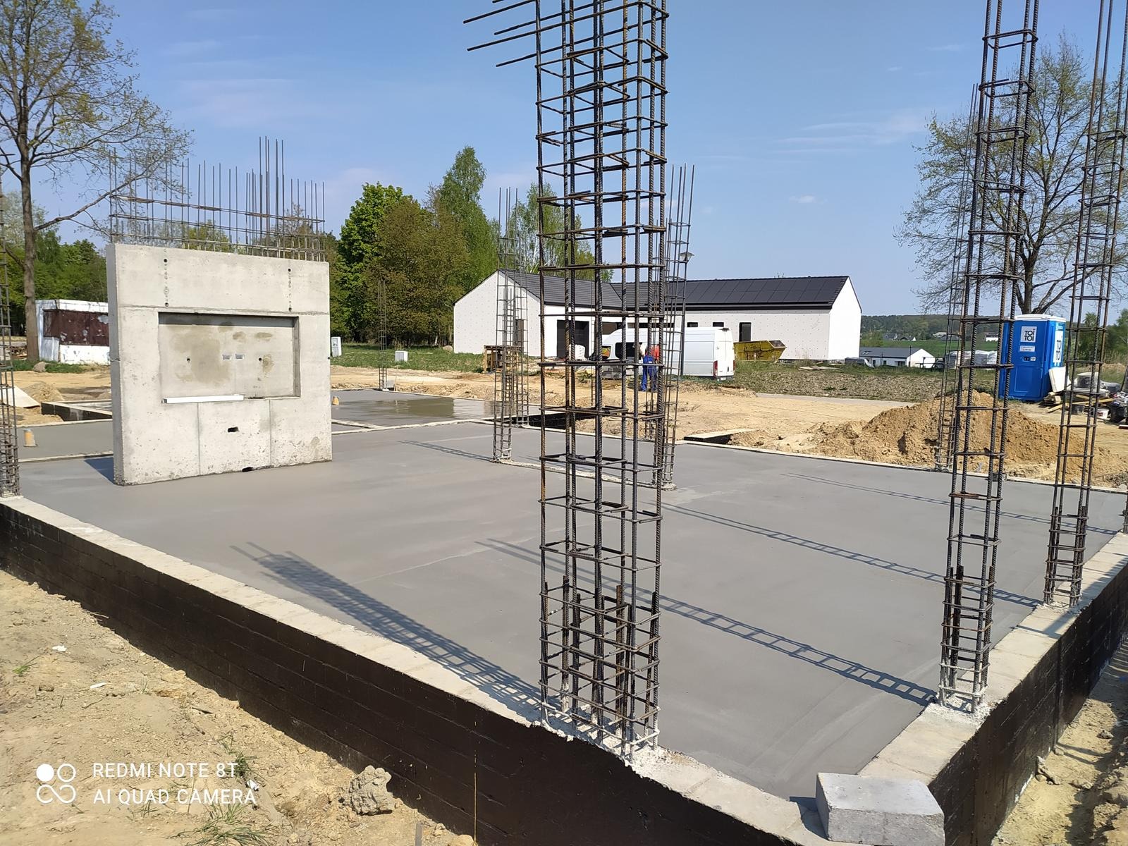 Zalany fundament z wystającymi prętami zbrojeniowymi, widoczny betonowy element konstrukcyjny. W tle budynek z panelami słonecznymi i przenośna toaleta.
