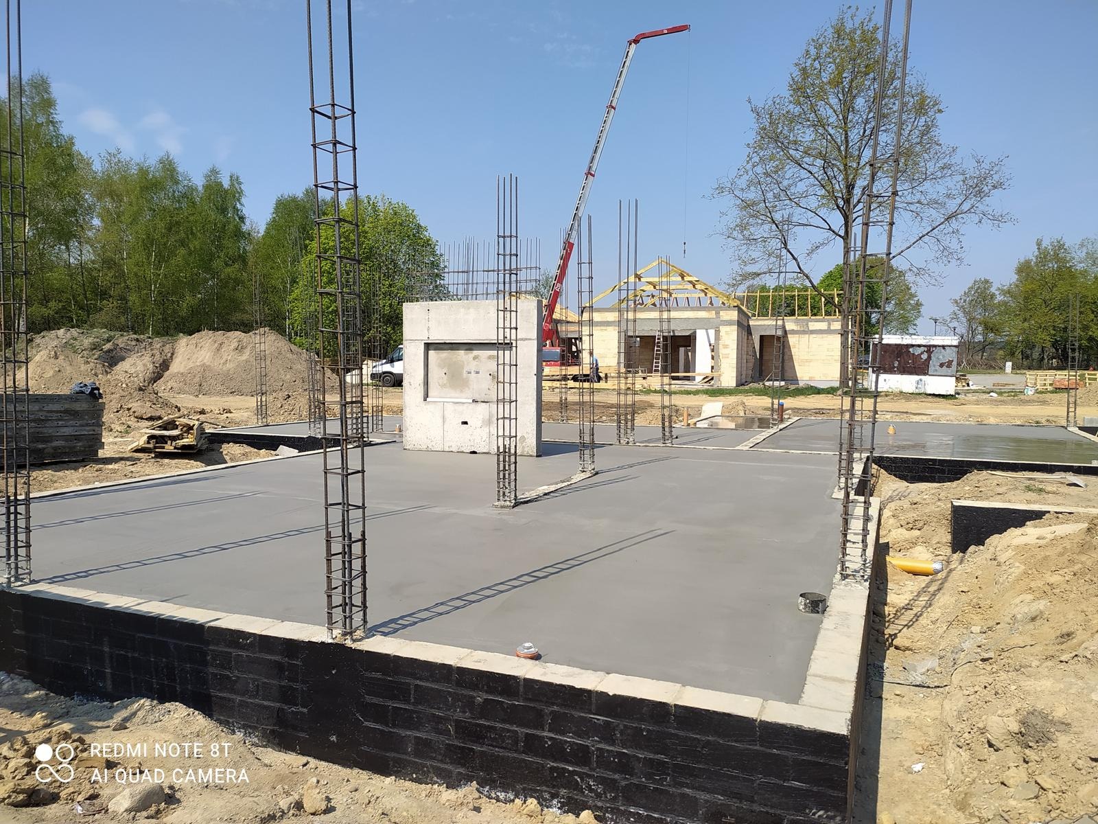 Wylany fundament z wystającymi prętami zbrojeniowymi na placu budowy domu jednorodzinnego. W tle widoczny dźwig i częściowo zadaszona konstrukcja budynku.