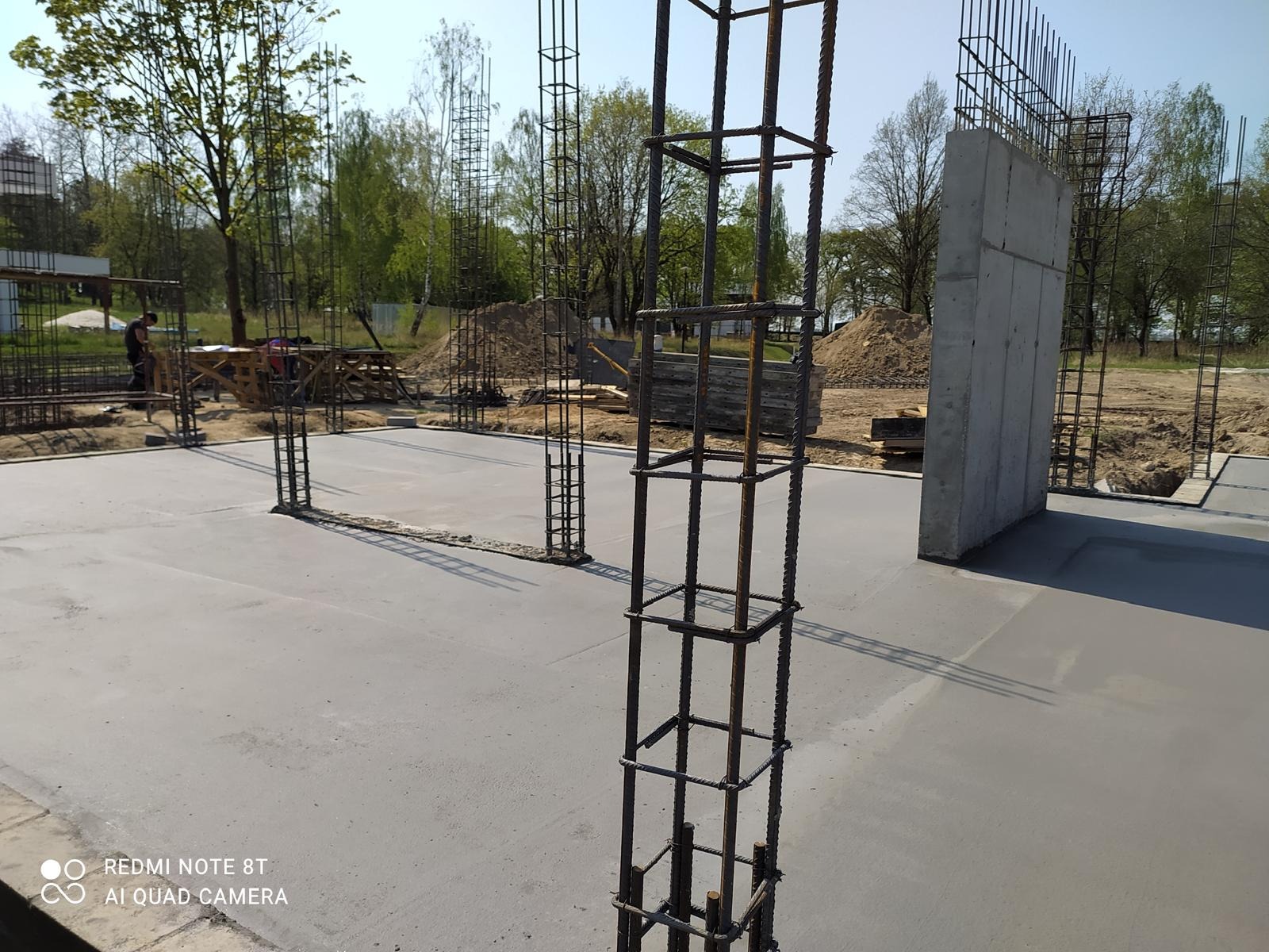 Betonowa płyta fundamentowa z wystającymi zbrojeniami kolumn i ścian. Widoczne ślady szalunków. W tle drzewa i hałdy ziemi.