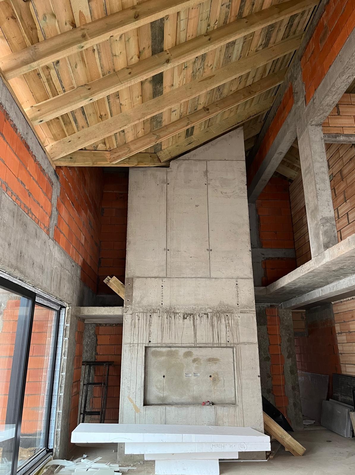 Wnętrze w trakcie budowy: surowy betonowy komin z wnęką, ceglane ściany, drewniany strop. Widoczne ślady po zalewaniu betonu, styropian na podłodze.