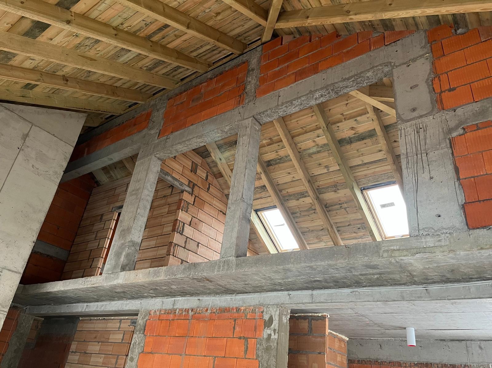 Wnętrze w trakcie budowy: surowy strop z drewnianymi belkami, fragmenty ścian z czerwonej cegły i betonowe filary. Widoczne okna dachowe i elementy konstrukcyjne.