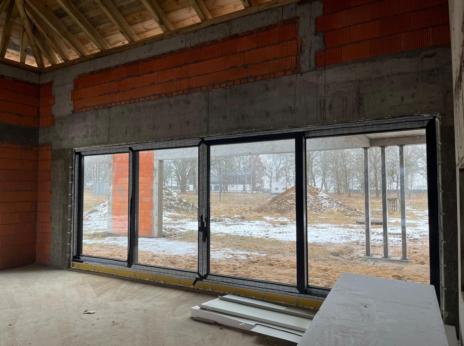Wnętrze surowego domu w trakcie budowy: ściany z betonu i cegły, duże okno przesuwne z widokiem na zaśnieżony krajobraz. Widoczne elementy konstrukcyjne dachu.