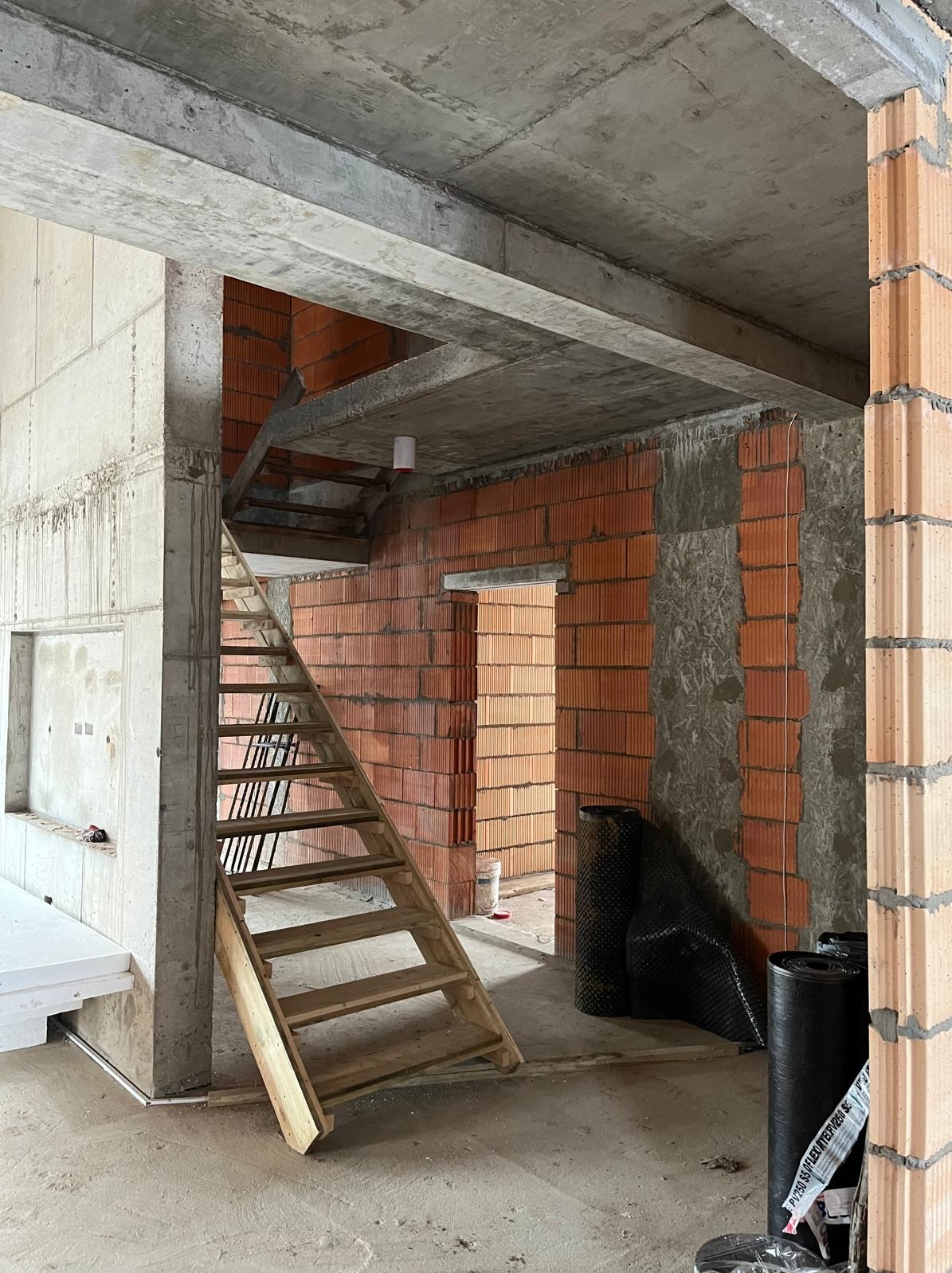 Wnętrze w trakcie budowy: ściany z czerwonej cegły, betonowy strop, drewniane schody tymczasowe. Widoczne materiały izolacyjne i surowy stan wykończenia.
