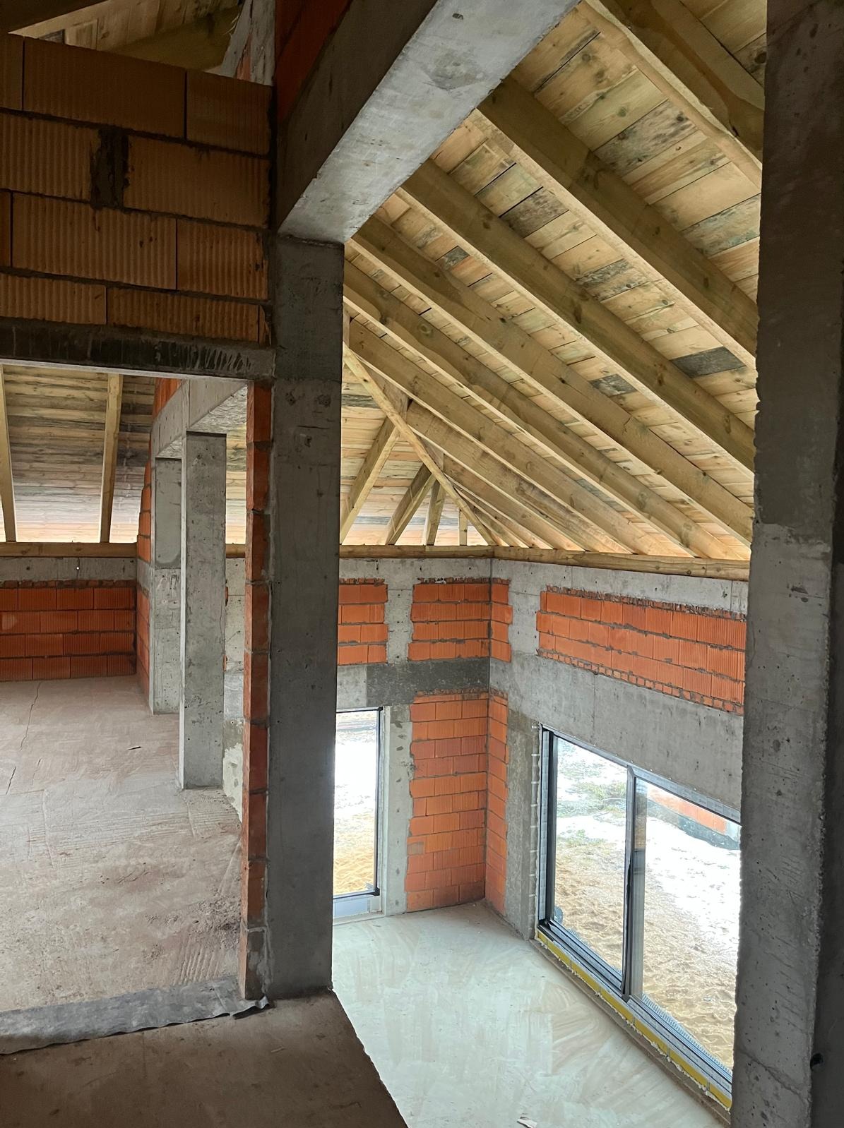 Wnętrze surowego domu w trakcie budowy: ściany z czerwonej cegły, betonowe filary i drewniany strop. Widoczne okna i surowa podłoga. Ujęcie z góry.