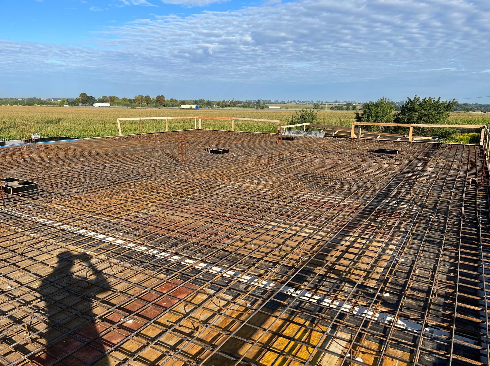 Zbrojenie płyty fundamentowej z widocznym szalunkiem i wystającymi prętami, na tle krajobrazu z polami i błękitnym niebem. Cień osoby w lewym dolnym rogu.