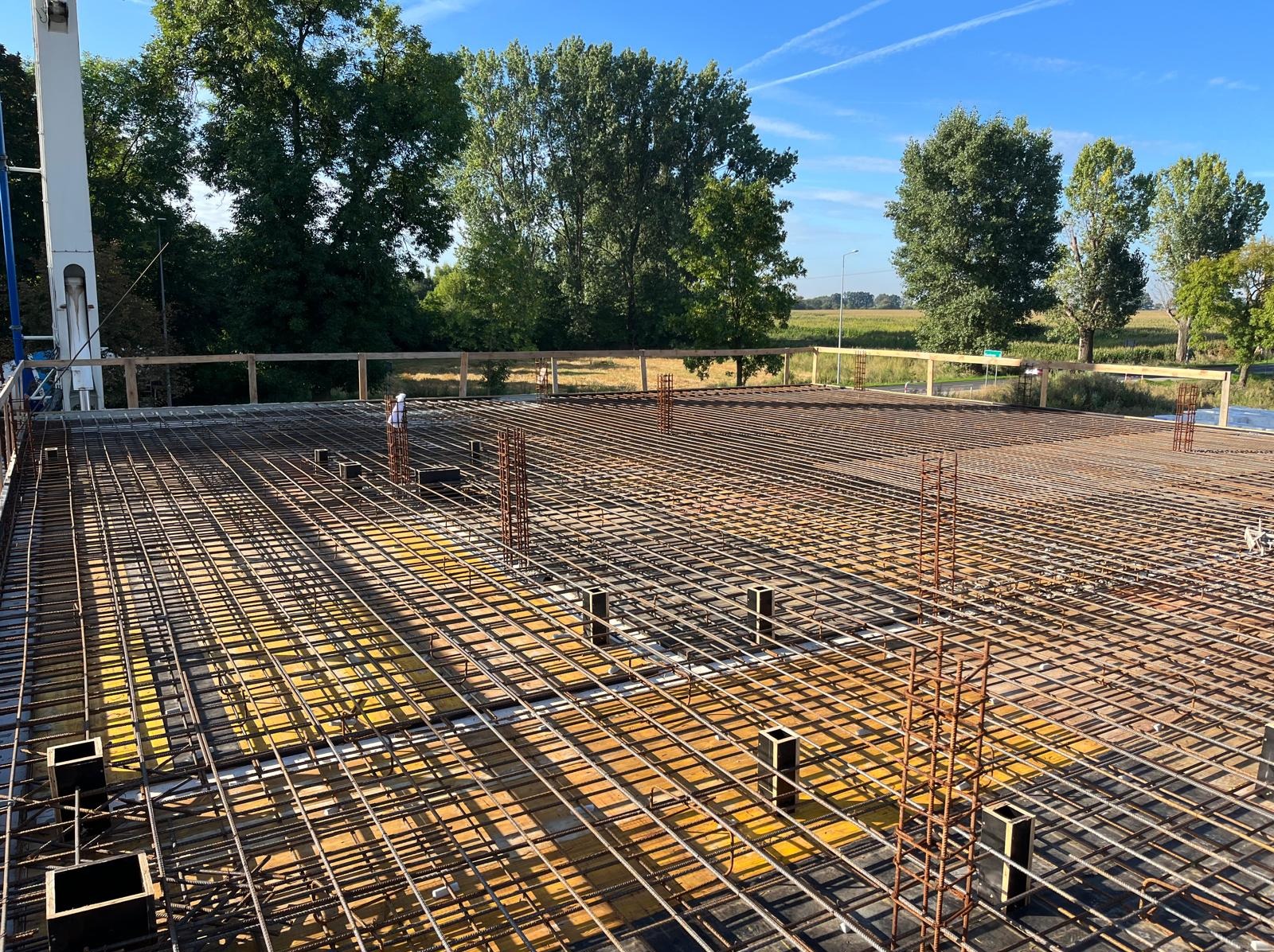 Zbrojenie płyty fundamentowej z widocznymi słupkami i siatką stalową na żółtej izolacji, w tle zieleń drzew i błękitne niebo. Prace budowlane w toku.