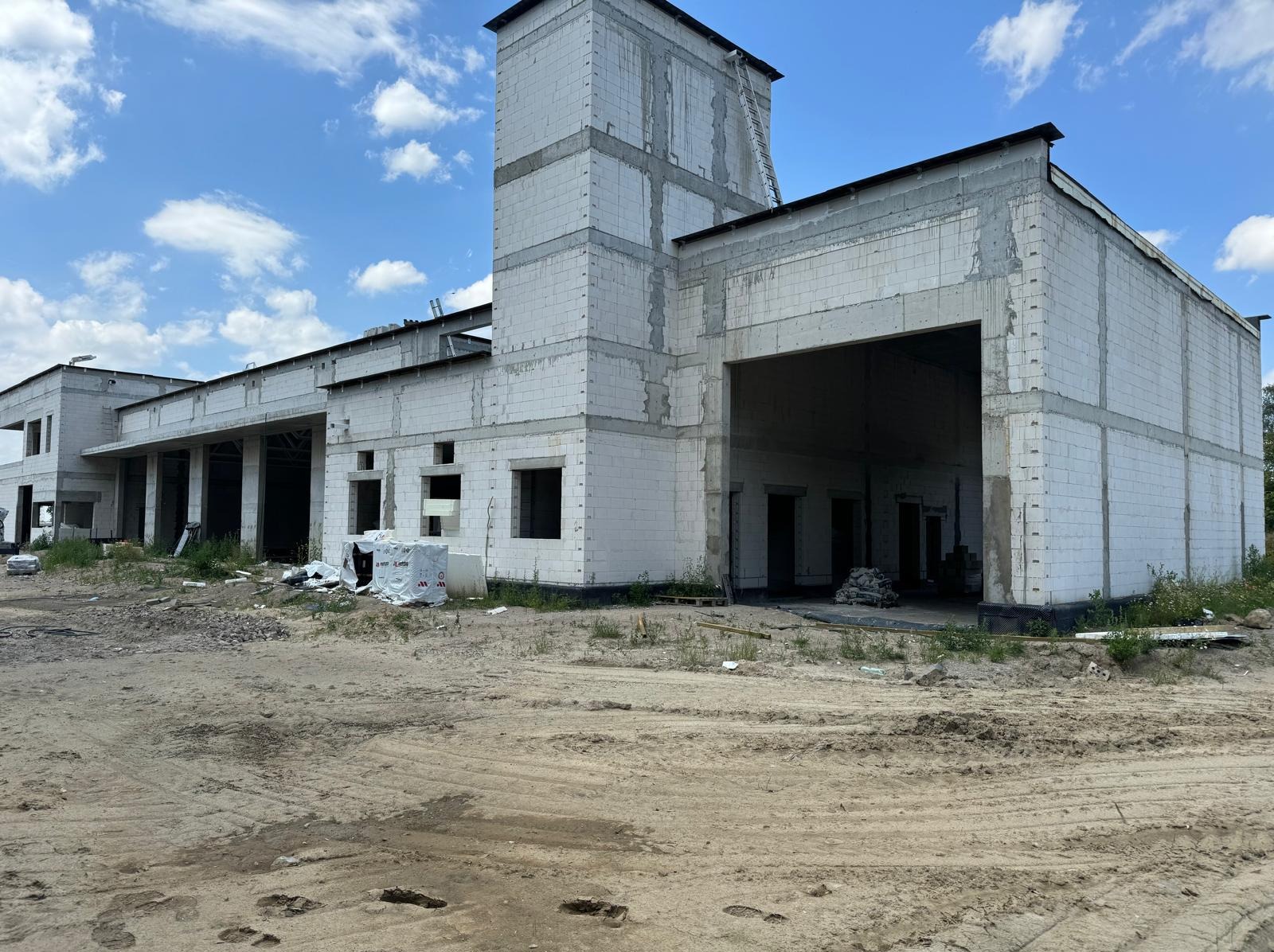 Budynek w stanie surowym otwartym, wzniesiony z białych bloczków, z widocznymi otworami okiennymi i bramą wjazdową. Teren wokół budynku piaszczysty, z widocznymi śladami opon.