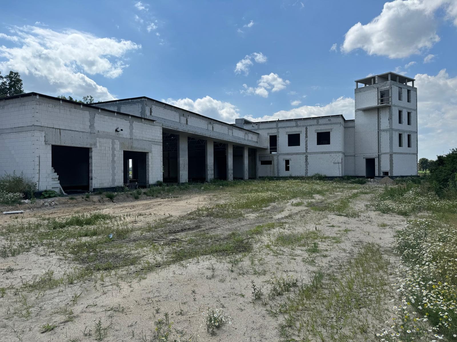 Niedokończony budynek z białego bloczka, z widoczną konstrukcją kolumnową i wieżą, otoczony piaszczystym terenem z roślinnością, pod błękitnym niebem z chmurami.