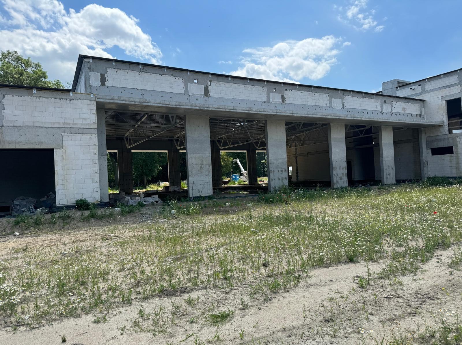 Surowy stan budynku z betonu komórkowego i żelbetu w trakcie budowy, widok z zewnątrz na tle błękitnego nieba z chmurami i zieleni. Widoczne kolumny i konstrukcja dachu.