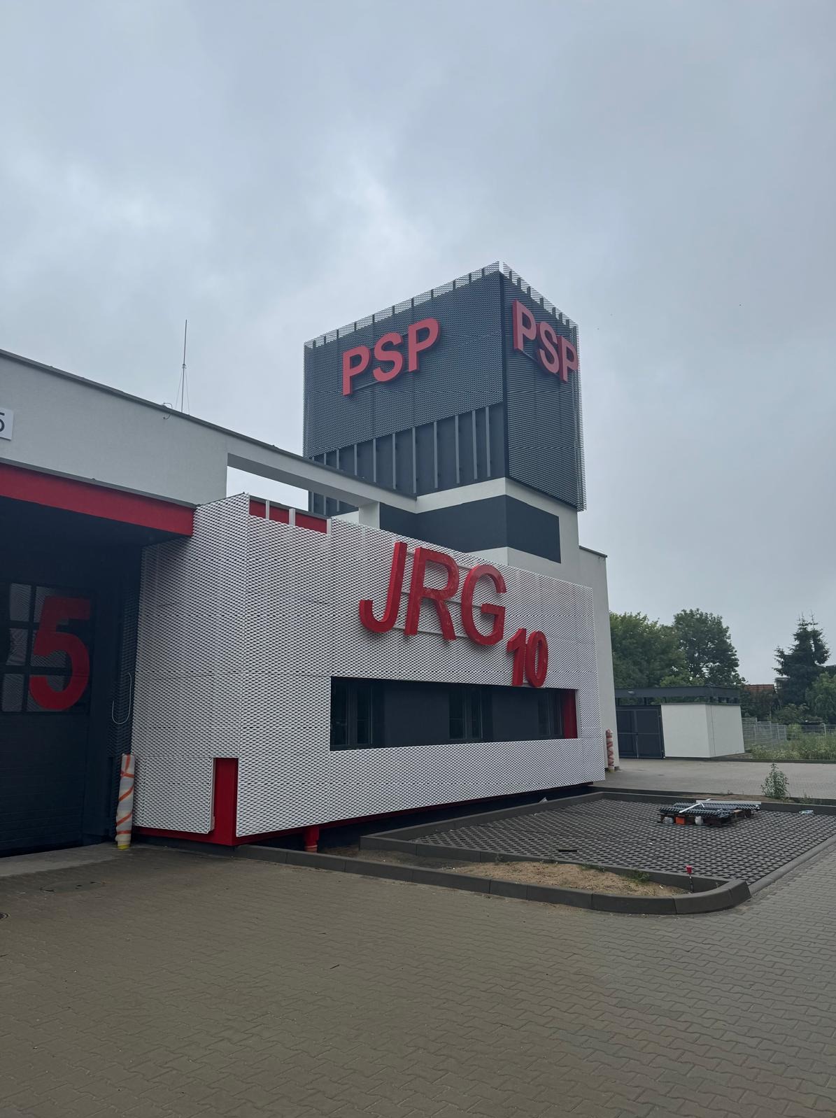 Nowoczesny budynek straży pożarnej z czerwonymi napisami PSP i JRG 10 na tle szarego nieba. Charakterystyczna elewacja z perforowanej blachy.