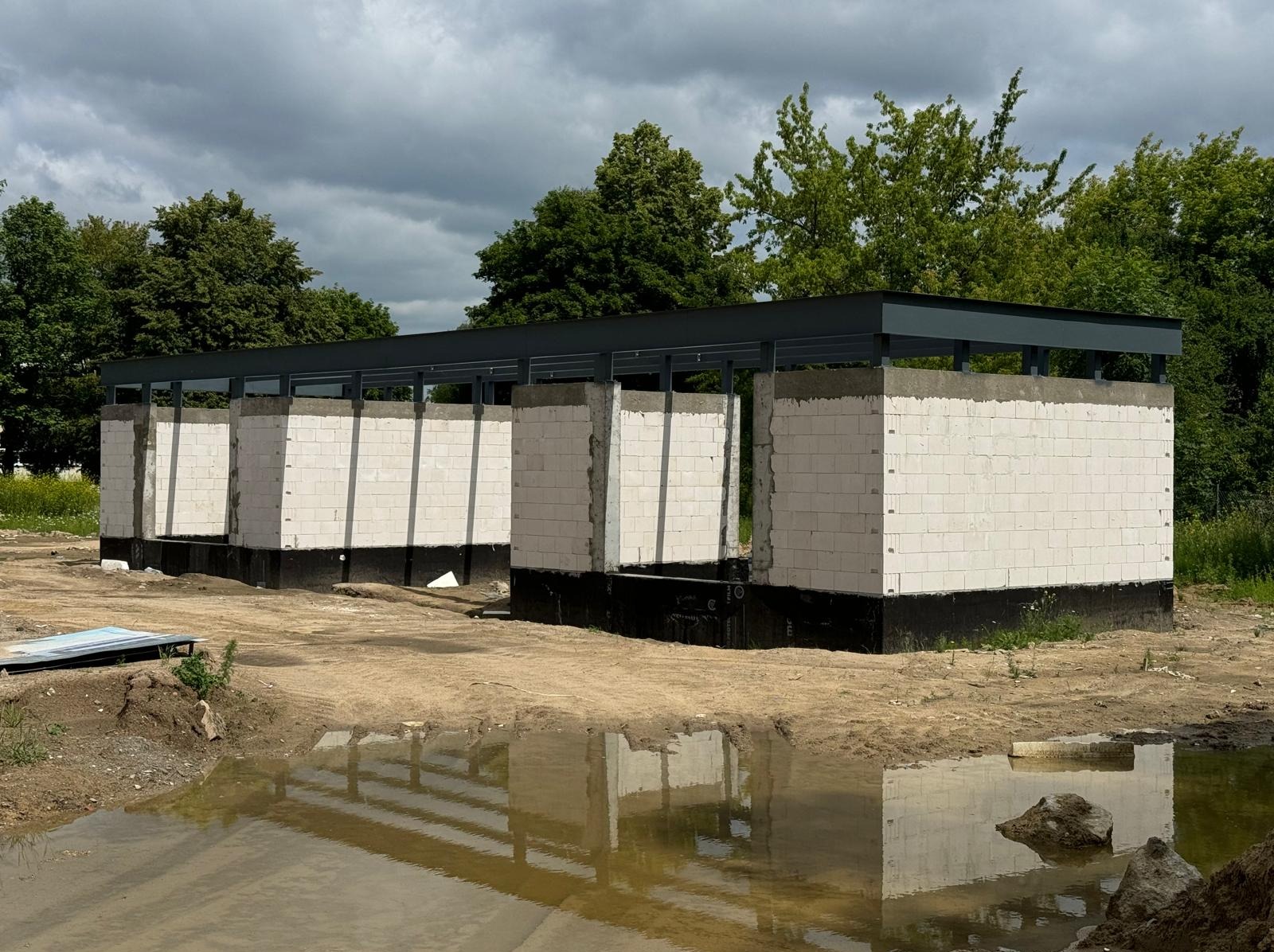 Wznoszenie ścian z białych bloczków na fundamencie z izolacją, z widoczną konstrukcją stalową dachu. W tle zieleń drzew i pochmurne niebo. Prace murarskie w toku.