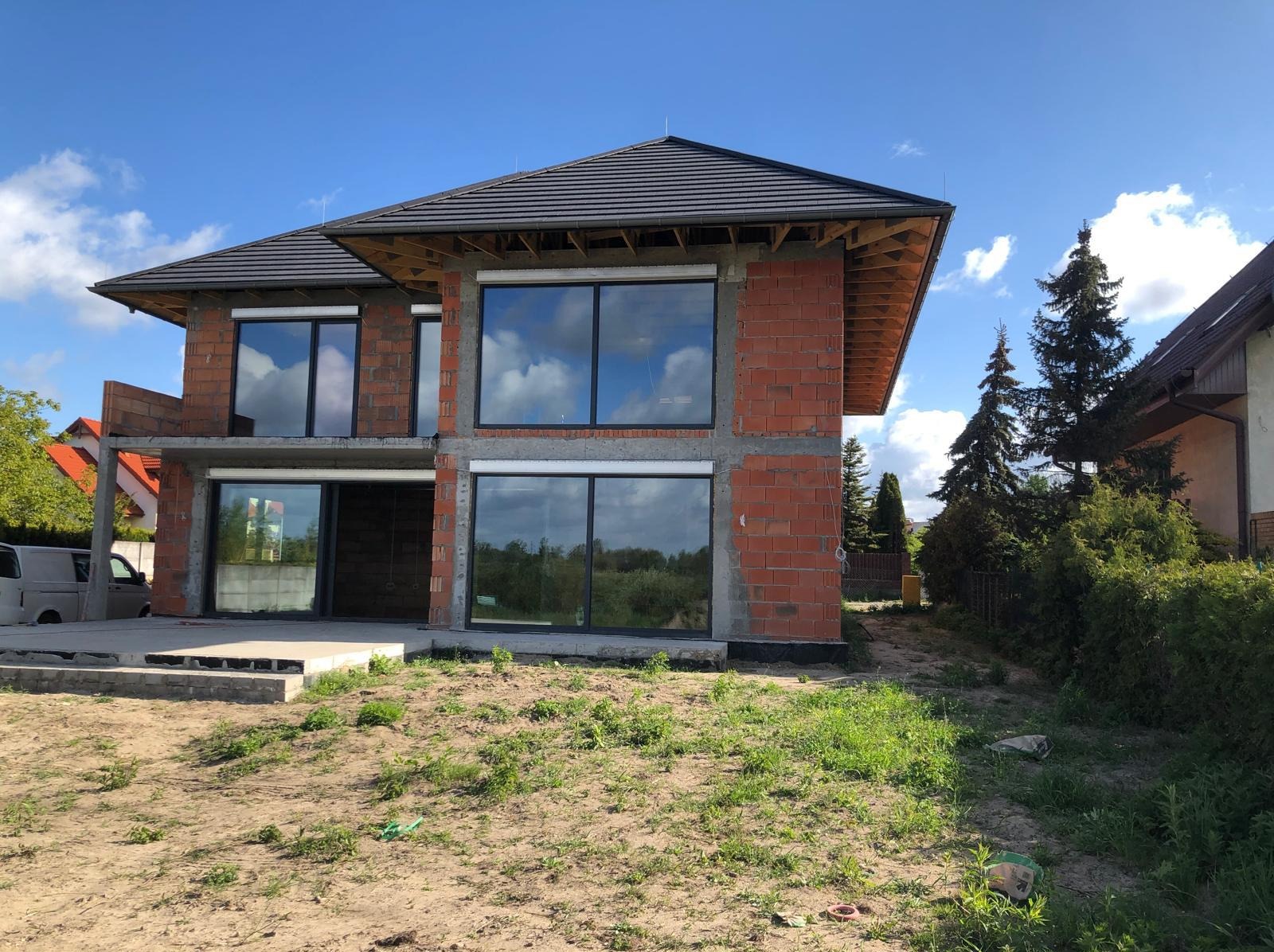 Dwukondygnacyjny dom w stanie surowym otwartym z dużymi oknami, widoczne cegły i betonowe elementy konstrukcyjne, dach pokryty dachówką. W tle błękitne niebo.