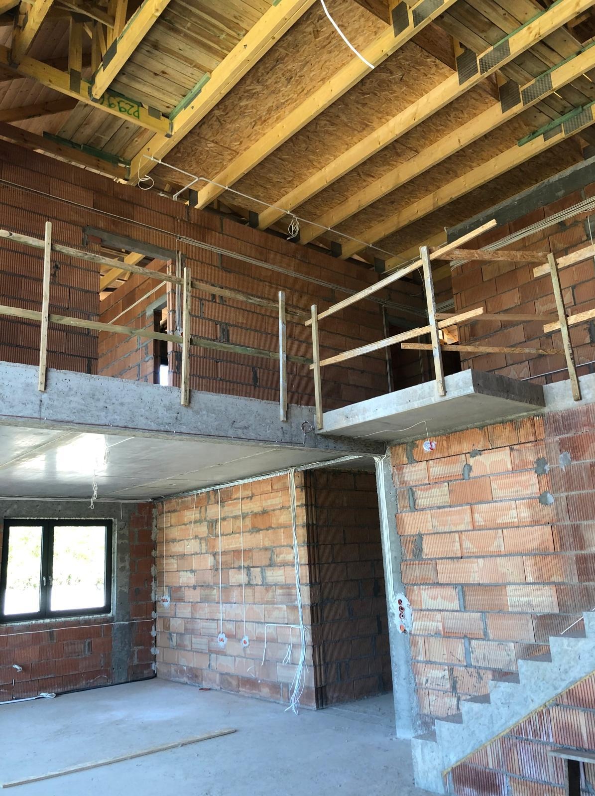 Wnętrze domu w trakcie budowy: ceglane ściany, betonowy strop, drewniana konstrukcja dachu, widoczne instalacje elektryczne. Surowy stan, okno z czarną ramą.