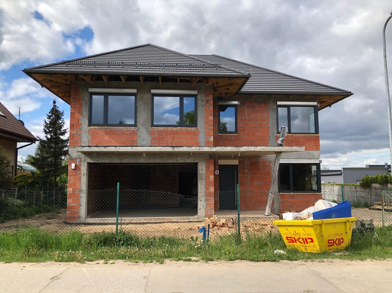 Dwukondygnacyjny dom w stanie surowym otwartym z cegły, z zamontowanymi oknami i szarą dachówką. Żółty kontener na gruz przed budynkiem.