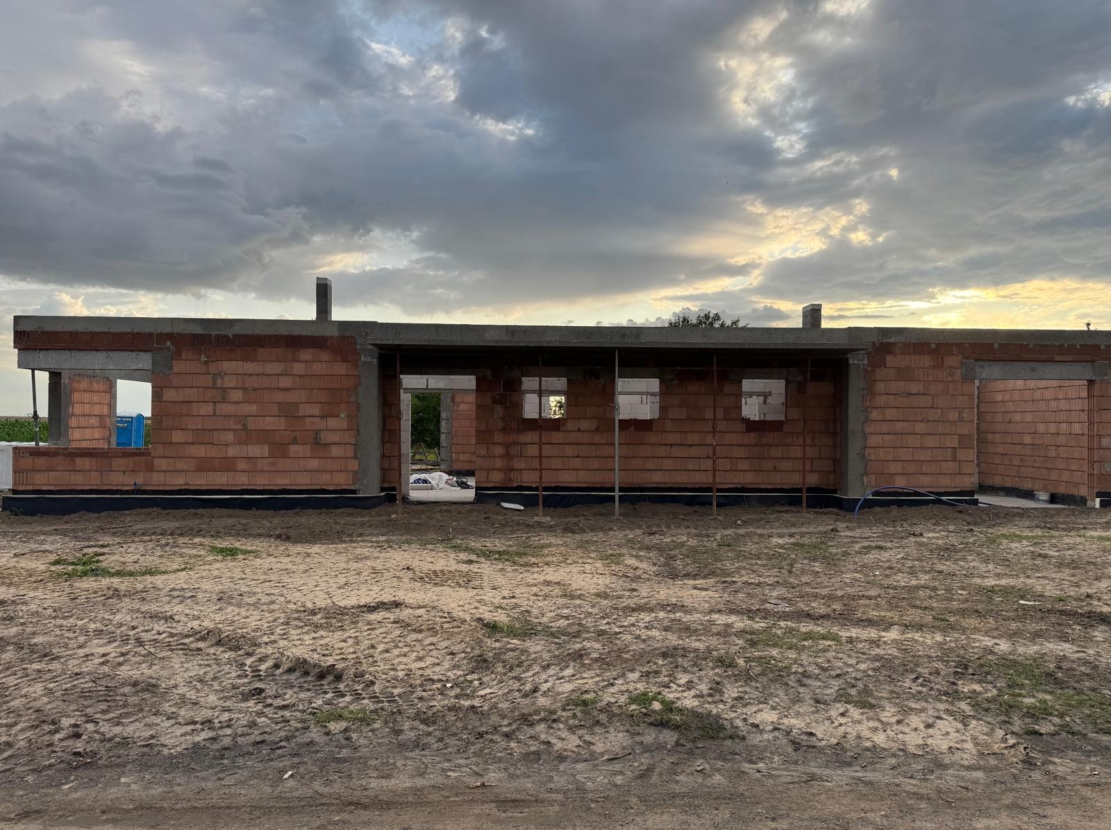 Wznoszenie ścian parterowego domu z czerwonej cegły. Widoczne otwory okienne i drzwiowe, fundamenty zabezpieczone czarną izolacją. Pochmurne niebo w tle.
