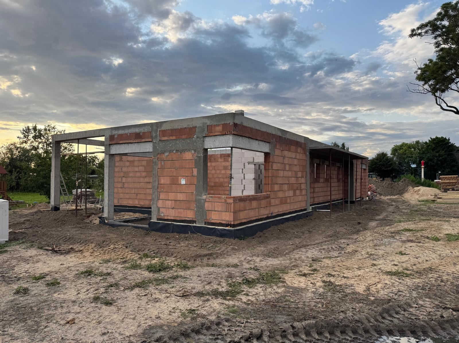 Surowy stan domu z czerwonej cegły i betonu pod pochmurnym niebem. Widoczne zbrojenia i pustaki. Etap budowy z widocznymi elementami konstrukcyjnymi.