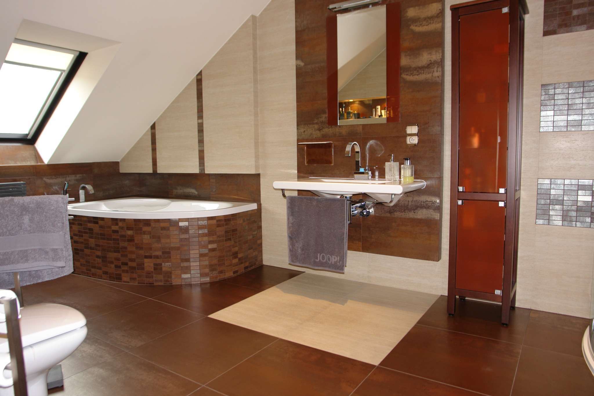 Nowoczesna łazienka na poddaszu z brązowymi płytkami, wanną obudowaną mozaiką, umywalką i wysoką szafką. Uwagę zwraca okno dachowe wpuszczające naturalne światło.